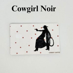 Pu&ograve; includere: Carta rettangolare con sfondo bianco e stelle rosse. Silhouette nera di una cowgirl che tiene una pistola, con il testo "Cowgirl Noir" in alto e "COWBOY CARTER" in basso.
