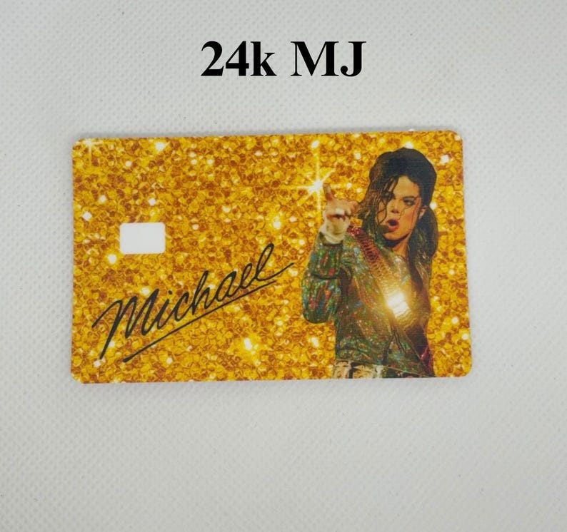 Pu&ograve; includere: Una carta color oro con uno sfondo glitterato e la scritta "24k MJ". La carta presenta una foto di Michael Jackson e il nome "Michael" in corsivo. La carta ha un ritaglio rettangolare bianco.