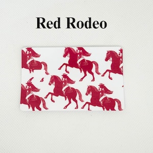 Pu&ograve; includere: Carta rettangolare bianca con un motivo rosso a tema rodeo ripetuto. Il motivo presenta sagome di una persona che cavalca un cavallo. Il testo "Red Rodeo" &egrave; stampato in nero nella parte superiore della carta.