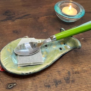 May include: A ceramic spoon rest with a green and blue glaze, holding a silver spoon with a green handle and two tea bags. A small teal glass candle holder with a lit candle sits in the background. The spoon rest is on a wooden surface.