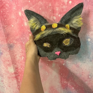 Peut inclure: Un masque de chat gris avec des détails jaunes, notamment des touffes d'oreilles et des cercles décoratifs. Le masque a un nez rose et est présenté sur un fond coloré sur le thème de la galaxie.