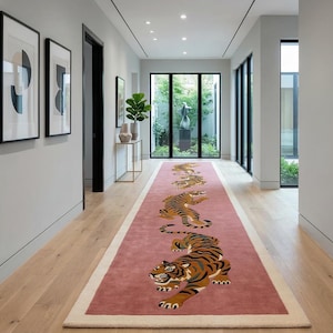 以下が含まれることがあります： ダスティローズピンクの背景に、オレンジ、黒、白の色合いで描かれた3頭の様式化された虎の繰り返し模様が特徴の、長方形の長いランナーラグ。ラグには無地のクリーム色の縁取りがあり、明るい木製の床の上に敷かれています。
