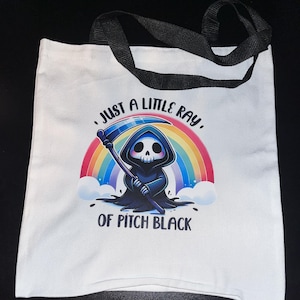 May include: White tote bag with black handles featuring a cartoon Grim Reaper character holding a scythe in front of a rainbow. The text reads "Just a little ray of pitch black."