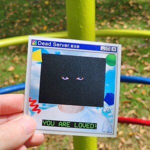 May include: A sticker with a retro computer interface design. The sticker features the text "Dead Server.exe" at the top and "YOU ARE LOVED!" at the bottom. The central graphic is a black box with two red eyes.