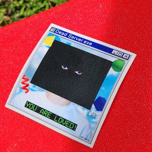 May include: A sticker with a retro computer window design. The top of the sticker reads "Dead Server.exe" in a blue bar. A black square covers the center, with two red eyes visible. The bottom of the sticker reads "YOU ARE LOVED!"
