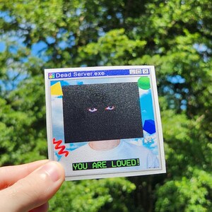 May include: A square sticker with a retro computer-style design. The sticker features the text "Dead Server.exe" at the top and "YOU ARE LOVED!" at the bottom. The center of the sticker has a black box with two eyes.