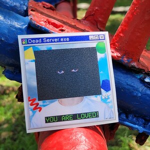 May include: A sticker with a retro computer window design. The text "Dead Server.exe" is at the top, and "YOU ARE LOVED!" is at the bottom. The sticker has a black square with two eyes.