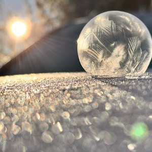 May include: A close-up shot of a frozen soap bubble with intricate ice crystal patterns. The bubble sits on a frosty surface, with the sun shining brightly in the background, creating a sparkling effect. The image captures the beauty of winter.