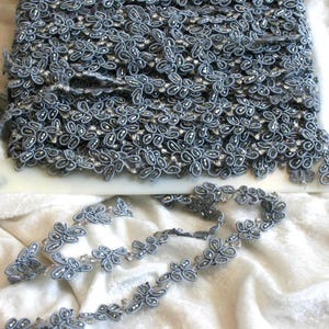 May include: A roll of gray floral lace with silver accents. The lace is intricately designed with small, flower-like patterns and is suitable for various craft projects. The lace is displayed on a white surface.