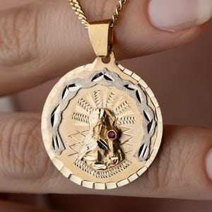 May include: A gold pendant necklace featuring a circular medallion with a central embossed figure of the Virgin Mary. The medallion has a textured gold background, a silver-toned outer rim with a repeating pattern, and a small red stone accent.