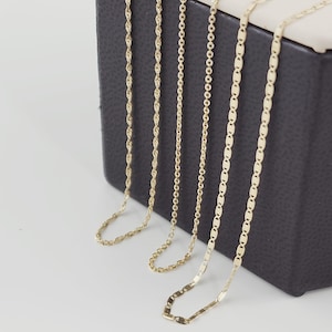 May include: Five gold necklaces with different chain styles are displayed against a dark gray background. The chains include rope, cable, and flat link designs. The necklaces are presented on a white surface, showcasing their varying textures and shine.