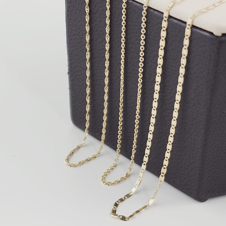 May include: Five gold necklaces with different chain styles are displayed against a dark gray background. The chains include rope, cable, and flat link designs. The necklaces are presented on a white surface, showcasing their varying textures and shine.