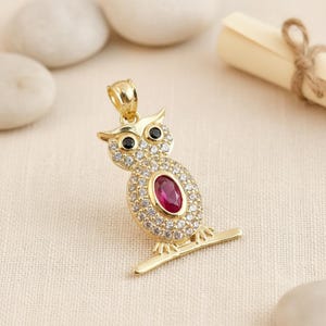 Può includere: Ciondolo a forma di gufo dorato con una pietra centrale color rubino e occhi neri. Il gufo è incastonato con piccole pietre trasparenti. Il ciondolo è su una barra dorata, su uno sfondo di tessuto beige chiaro.