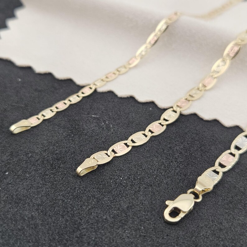 May include: A tri-color gold chain necklace featuring alternating oval links in yellow, white, and rose gold tones. The links have a textured, starburst-like pattern on their surfaces. The chain includes a secure lobster claw clasp closure.