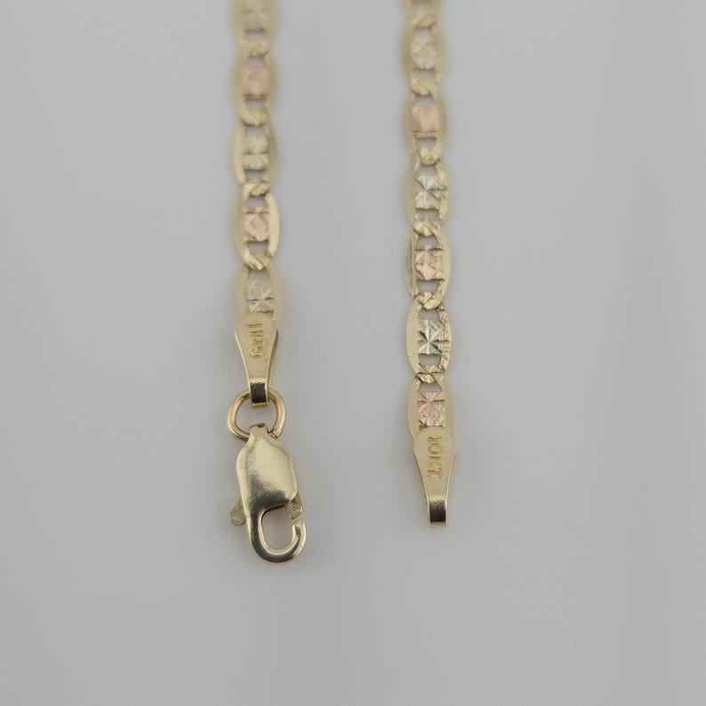May include: A gold chain necklace featuring alternating oval links with a textured, faceted finish and smooth rose gold accents. The chain includes a lobster claw clasp and a metal tag stamped with the text 10KT, indicating the gold purity.