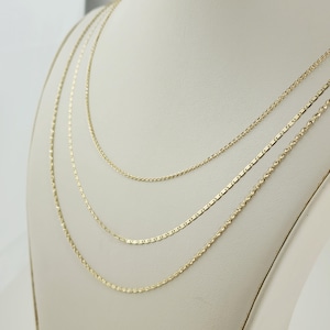 May include: Three delicate gold necklaces displayed on a white bust. The necklaces feature different chain styles: a thin rope chain, a flat rectangular chain, and a delicate cable chain. The chains are all a warm gold color.