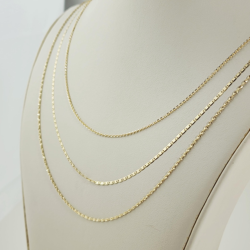 May include: Three delicate gold necklaces displayed on a white bust. The necklaces feature different chain styles: a thin rope chain, a flat rectangular chain, and a delicate cable chain. The chains are all a warm gold color.