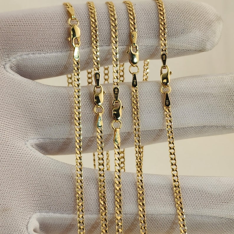 May include: Close-up of several gold-colored curb chain necklaces. Each chain has a lobster clasp closure. The chains are displayed against a white background. The chains are likely made of gold.