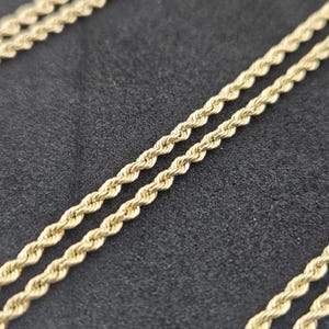Op de afbeelding: Een goudkleurige touwketting met een gedraaid, spiraalvormig schakelontwerp. De ketting bestaat uit meerdere strengen van onderling verbonden metalen schakels die een doorlopend, getextureerd en uniform uiterlijk creëren over de gehele lengte.