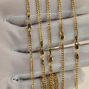 May include: Close-up of multiple gold-colored curb chain necklaces. Each chain is connected with a lobster clasp. The chains are displayed on a light gray surface. The chains are likely made of gold.