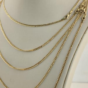 May include: Several gold-colored curb chain necklaces displayed on a white mannequin. The chains vary in thickness and length, each with a lobster clasp closure. The necklaces are presented against a neutral background, highlighting their shiny, metallic appearance.