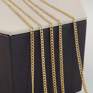 May include: Close-up of multiple gold-colored curb chains. The chains are displayed vertically, showcasing their linked design. The chains are on a dark gray surface with a light gray top.