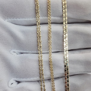 May include: Close-up of three gold necklaces with different chain styles. The chains are displayed on a white fabric background. The necklaces have a shiny, reflective surface.