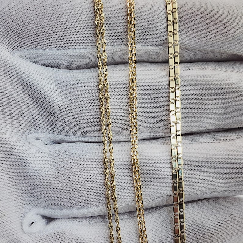 May include: Close-up of three gold necklaces with different chain styles. The chains are displayed on a white fabric background. The necklaces have a shiny, reflective surface.
