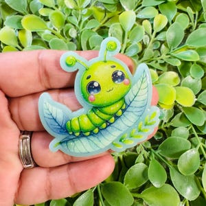 May include: A die-cut sticker featuring a bright green cartoon caterpillar with large, dark, starry eyes and pink cheeks. The caterpillar is resting on a light blue leaf with a subtle glittery finish and a small, curved antenna on its head.