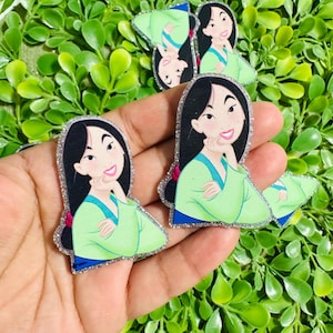 May include: Several glittery resin embellishments featuring a cartoon character in a green dress with black hair. The character is smiling and has a hand under their chin. The embellishments are surrounded by green foliage.