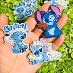 May include: Three acrylic pins featuring the character Stitch. One pin shows Stitch with the text Stitch and a heart. Another shows Stitch hugging a brown teddy bear with a red heart. The third shows Stitch with three white ducks and a doll.