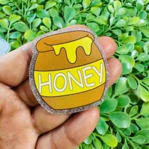 May include: A honey pot-shaped charm with a brown body and yellow honey dripping from the top. The word "HONEY" is displayed in white on a yellow banner across the pot. The charm has a glittery, clear border.