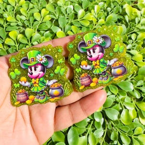 May include: Two glittery green Minnie Mouse-themed St. Patrick's Day decorations. Each features Minnie in a green outfit and hat, holding a gold coin, with a pot of gold and shamrocks. The background is a bed of green leaves.