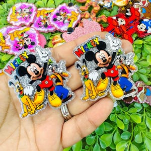 K&ouml;nnte beinhalten: Glitzernde Mickey Mouse-, Goofy- und Pluto-Charakterverzierungen. Mickey tr&auml;gt ein rotes Hemd, Goofy ein orange-gelbes Outfit und Pluto ist gelb. Das Wort "Mickey" ist rot. Weitere Charakterverzierungen befinden sich im Hintergrund.