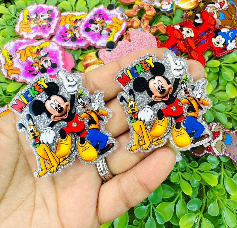 K&ouml;nnte beinhalten: Glitzernde Mickey Mouse-, Goofy- und Pluto-Charakterverzierungen. Mickey tr&auml;gt ein rotes Hemd, Goofy ein orange-gelbes Outfit und Pluto ist gelb. Das Wort "Mickey" ist rot. Weitere Charakterverzierungen befinden sich im Hintergrund.