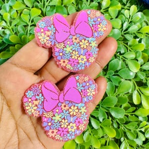 May include: Two pink Minnie Mouse-shaped embellishments with a floral design. The ears and bow are a solid pink, while the inside is filled with colorful flowers. The edges have a glittery pink border. These are likely for crafting or accessories.