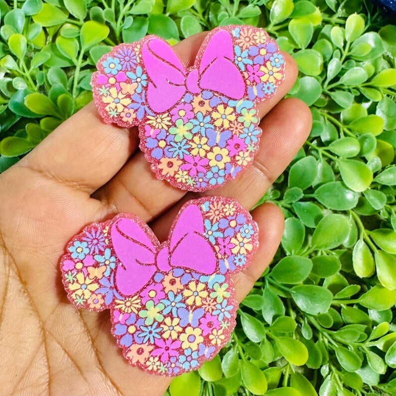 May include: Two pink Minnie Mouse-shaped embellishments with a floral design. The ears and bow are a solid pink, while the inside is filled with colorful flowers. The edges have a glittery pink border. These are likely for crafting or accessories.