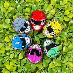 May include: A collection of six Power Rangers helmet pins featuring red, yellow, green, pink, blue, and grey designs. Each helmet is outlined with a glittery border and displays the distinct facial features and visor of the iconic character.