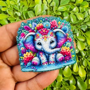 May include: A colorful, whimsical elephant design. The elephant is adorned with floral patterns and surrounded by vibrant flowers in shades of pink, blue, and yellow. The background is a glittery teal.