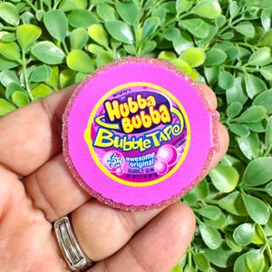 May include: A circular pink resin charm featuring the Hubba Bubba Bubble Tape logo. The design includes the text Wrigley's Hubba Bubba Bubble Tape, 6 Feet of Fun, and Awesome Original Bubble Gum. The edges are textured with a glittery pink finish.