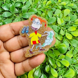 May include: A glittery acrylic pin featuring the cartoon character Eliza Thornberry with orange pigtails, wearing a yellow shirt and red shorts. The character holds blue binoculars in one hand and an open map in the other, set against a gold background.