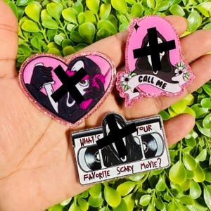 May include: Three glittery acrylic pins featuring a masked figure in a black hooded robe. One heart-shaped pin shows the figure holding a knife and phone. A tombstone-shaped pin says CALL ME. A rectangular VHS tape pin says WHAT'S YOUR FAVORITE SCARY MOVIE?