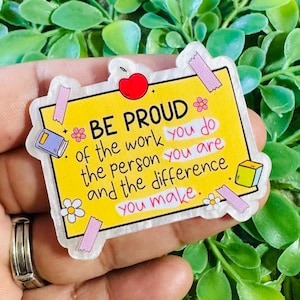 May include: A rectangular yellow sticker with a white border featuring the text BE PROUD of the work you do the person you are and the difference you make. The design includes a red apple, small flowers, and purple tape accents.