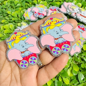 May include: A set of glittery acrylic charms featuring the character Dumbo, a light blue elephant with large pink ears, wearing a yellow hat and sitting inside a colorful circus wagon with red, blue, and green accents and a silver glitter border.