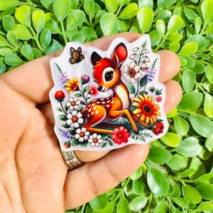 May include: A die-cut sticker featuring a cartoon fawn with large eyes and white spots, surrounded by colorful wildflowers, daisies, and a small butterfly. The illustration uses a vibrant palette of orange, red, yellow, and white against a white background.
