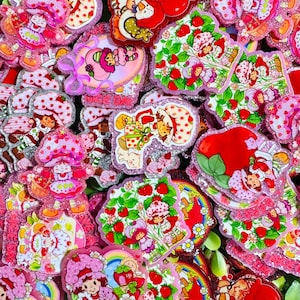 May include: A collection of glittery acrylic charms featuring Strawberry Shortcake characters, strawberries, and green leaves. The designs include various poses of the characters, some holding gifts or baskets, with vibrant red, pink, and green colors.
