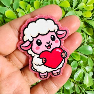 May include: A pink glittery sheep-shaped sticker holding a red heart. The sheep has white fluffy wool, pink ears, and a smiling face with black eyes. The sticker is held in a hand against a background of green leaves.