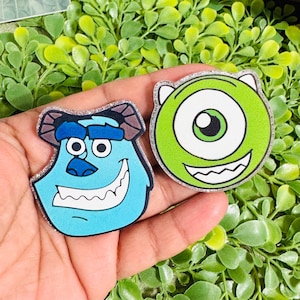 May include: Two circular acrylic pins featuring cartoon characters. One pin shows a blue, furry-textured creature with purple spots and horns. The other pin shows a round, green, one-eyed creature with a wide, toothy grin and small horns.