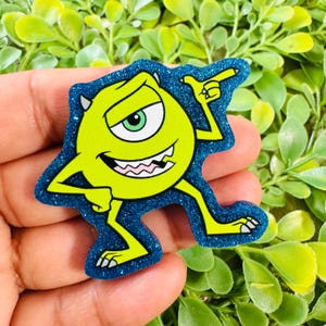 May include: A lime green, one-eyed monster character with two small horns, a wide grin, and a raised arm, set against a dark blue, glittery, die-cut background. The character has a single large eye with a green iris and sharp white teeth.