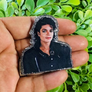 May include: A flat acrylic pin featuring a portrait of Michael Jackson with his signature curly dark hair, wearing a black collared shirt with visible silver button details. The pin has a clear, textured, glittery border edge.